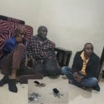 Men sitting on the floor during police arrest, with confiscated jewelry and phones below, in a room with minimal furniture, illustrating a news scene related to crime investigation in Uganda.